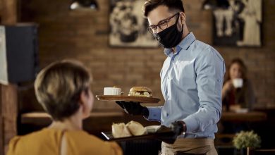 Happy waiter wearing protective face mask and gloves while bringing food to a customer in a pub.